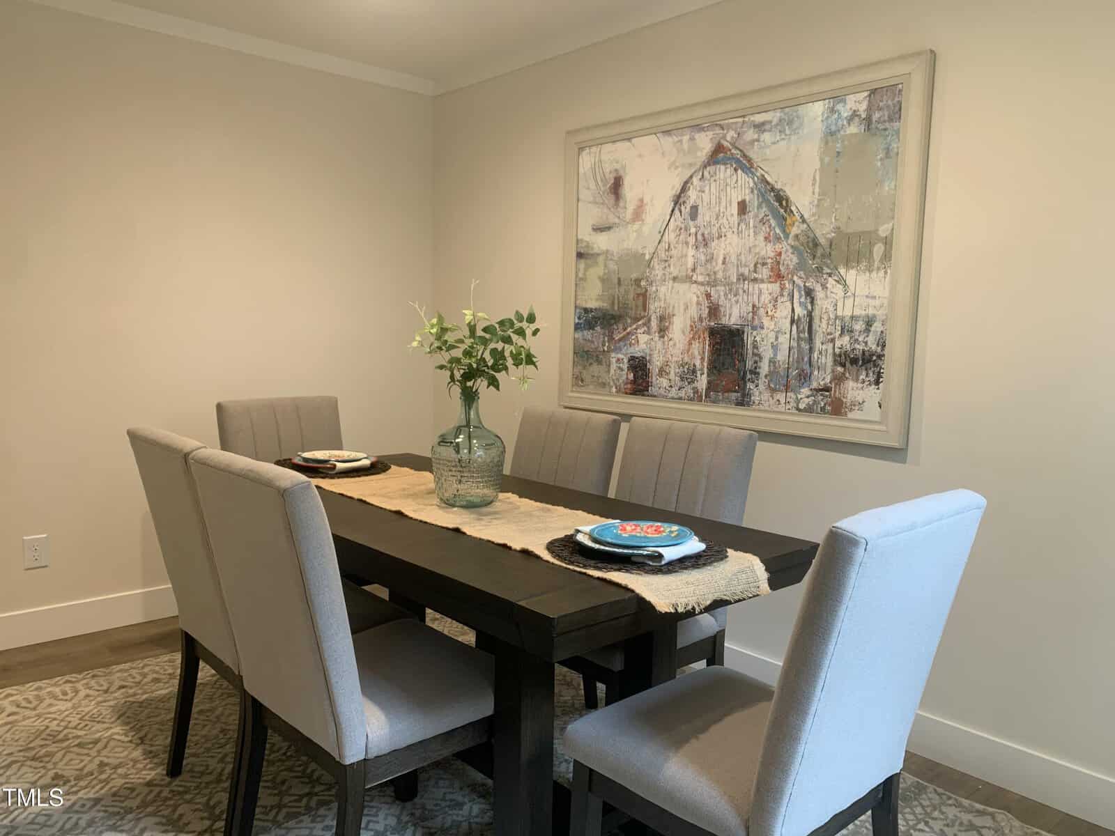 A dining area with a dark wood table, four cushioned gray chairs, a beige table runner, a vase with green leafy branches, and a large barn painting on a cream-colored wall.