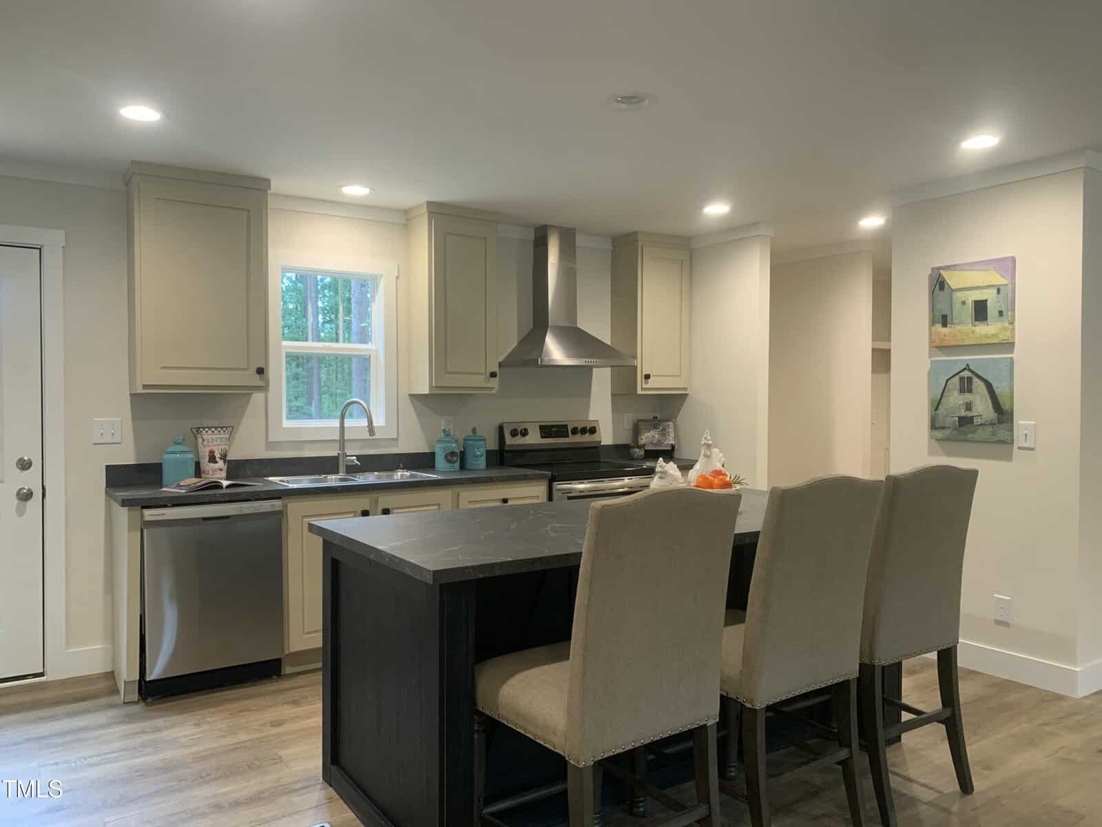 Modern kitchen with light tan cabinets, a stainless steel oven and dishwasher, a black countertop island with three beige upholstered chairs, and two paintings of houses on the wall. Wood flooring and recessed lighting are present.
