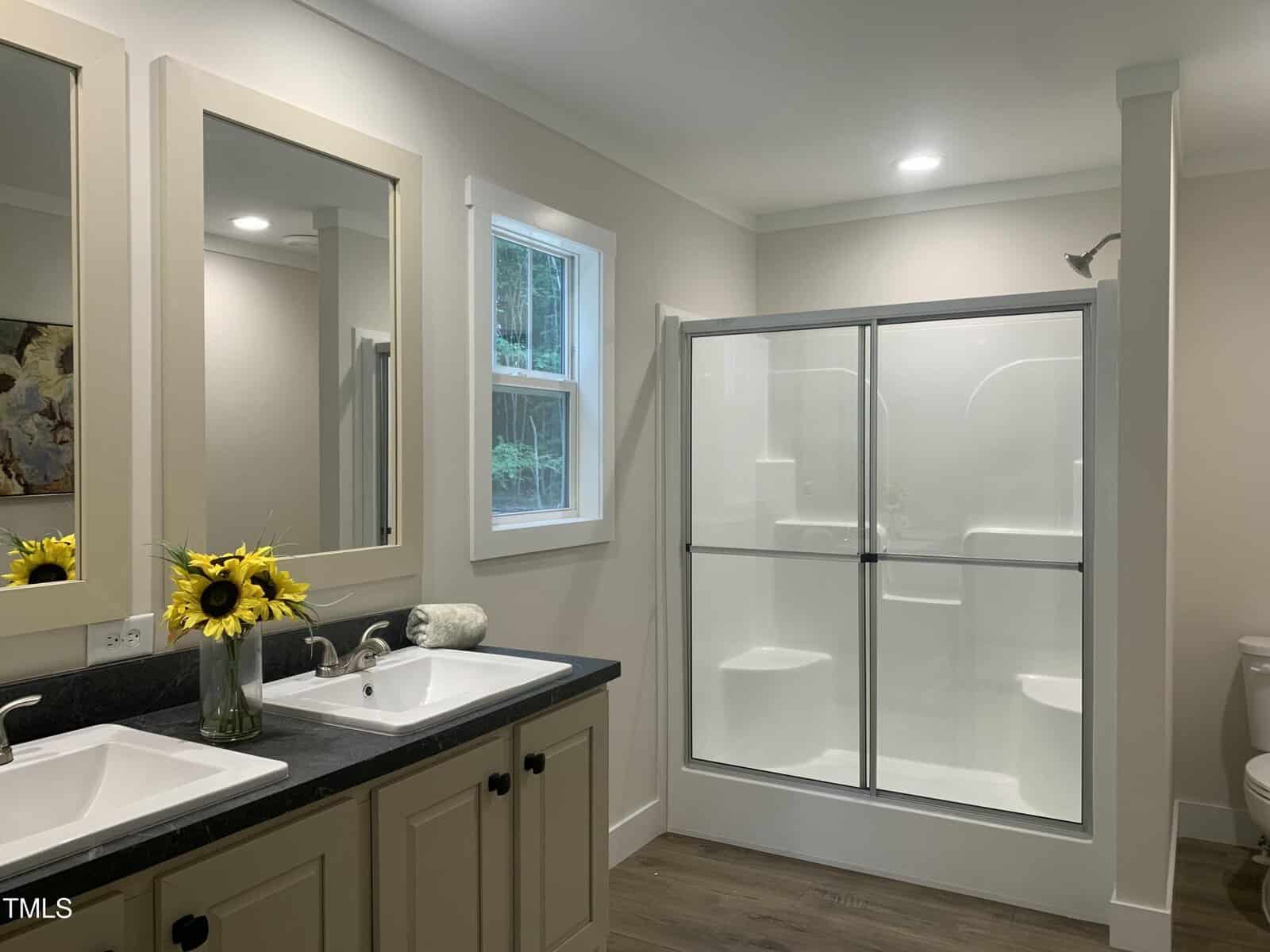 A modern bathroom with a double sink vanity, a vase of sunflowers, a window, a glass-enclosed shower, and a toilet. The room has light walls and wood-look flooring.