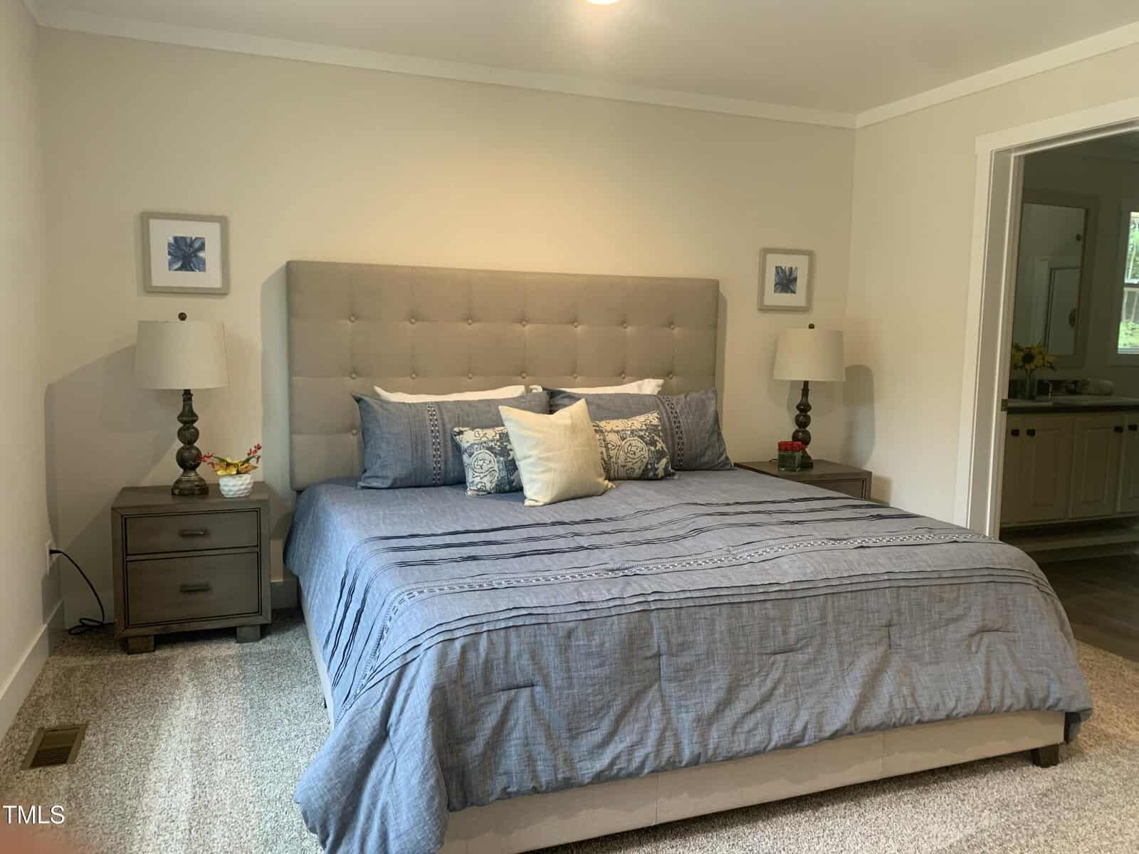 A bedroom with a large bed featuring a tufted beige headboard, blue and white bedding, two nightstands with matching lamps, framed art on the wall, and an open doorway leading to a bathroom.