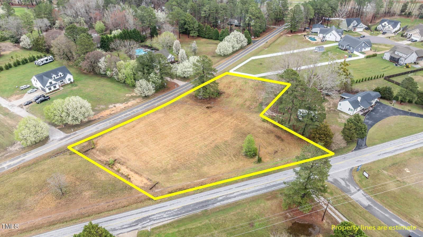 Aerial view of a cleared, grassy lot outlined in yellow at 0 Wiggins Rd Road, Louisburg, bordered by paved roads on two sides, with neighboring houses, driveways, and trees surrounding the property. Text states “Property lines are estimate.”.
