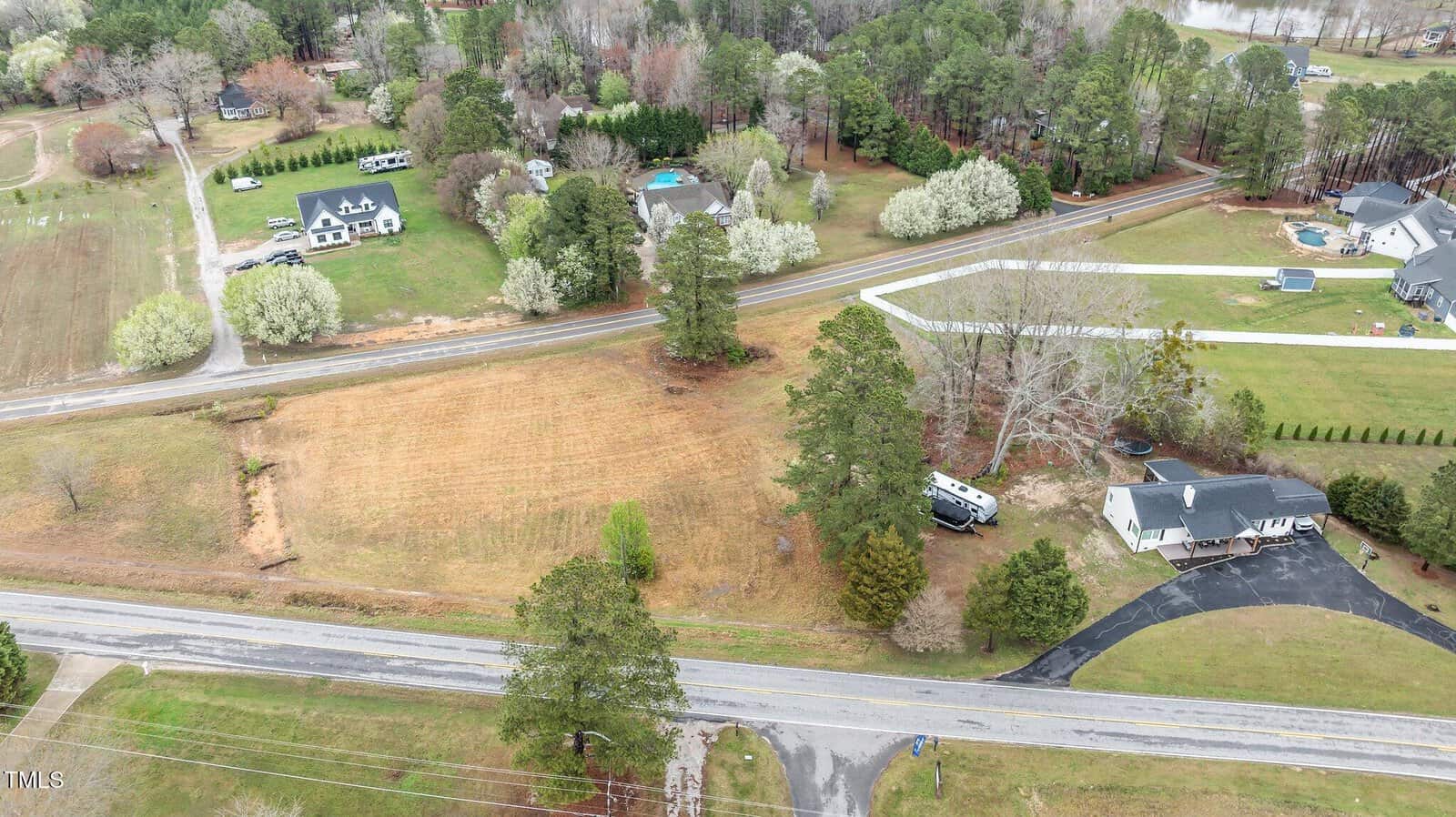 Aerial view of a rural area with a few houses, trees, a road running horizontally, a driveway, a lawn, and an RV parked near a house. Other houses and open fields are visible in the background.
