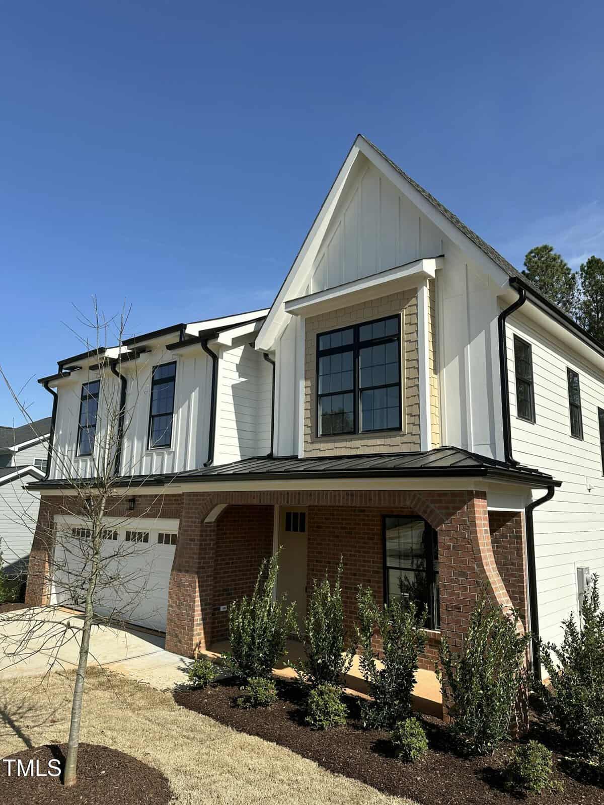 Two-story modern house with white siding, black trim, and large windows. The front features a brick archway over the porch, landscaped bushes, and a double garage. The sky is clear and blue.