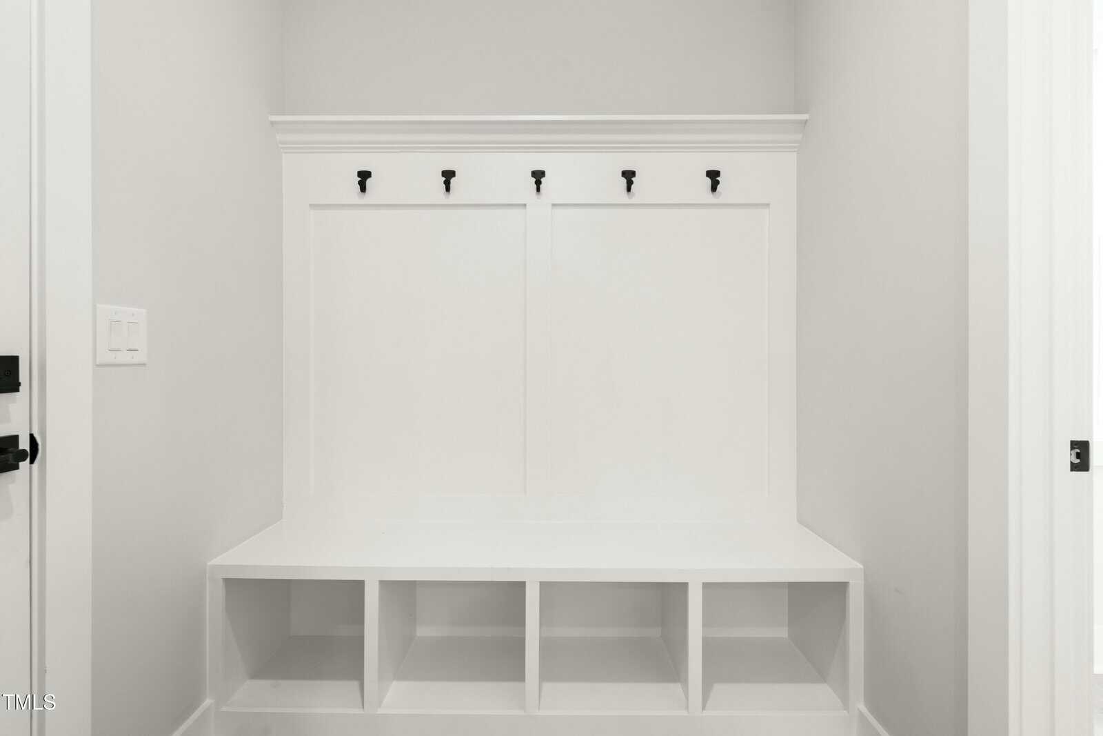 A white built-in mudroom bench with four cubby shelves underneath and five black hooks mounted above on a paneled white wall, set in a bright, minimalist space.