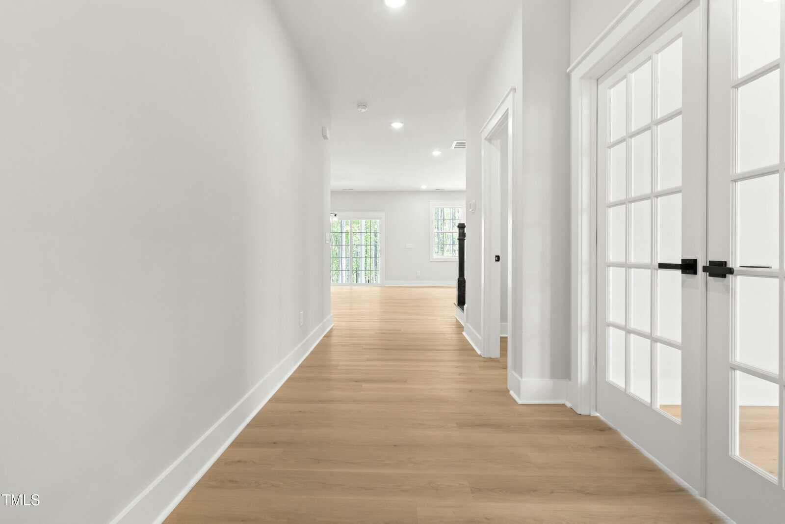 Bright hallway with light wood floors, white walls, and double glass doors on the right; the space opens into a sunlit room with large windows at the end.