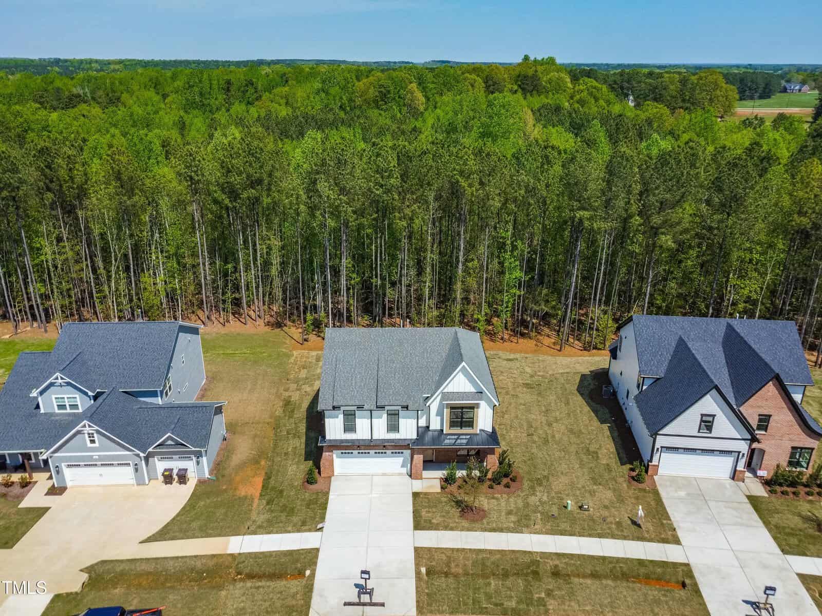 An aerial view of three modern suburban houses with driveways and neatly manicured lawns, bordered by a dense forest of tall green trees in the background.