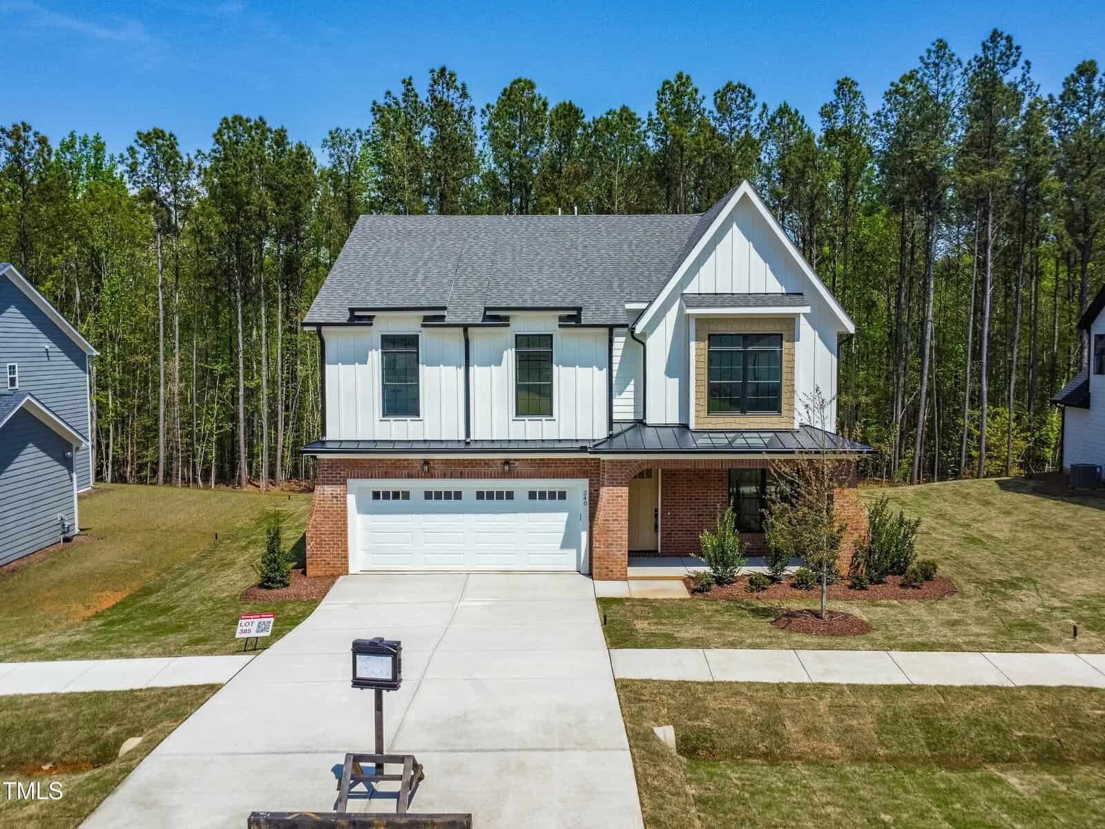 A modern two-story house with white siding and brick accents, a double garage, and a large driveway, set against a backdrop of tall trees and a neatly maintained lawn.