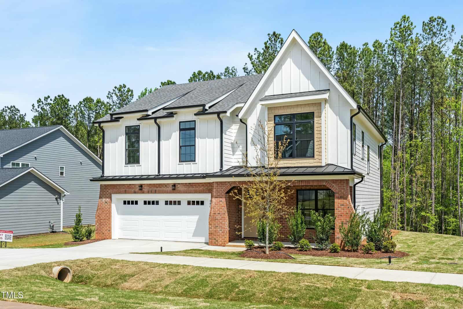 A modern two-story house with a combination of white siding and red brick exterior, large windows, a two-car garage, small front yard, and surrounded by trees under a clear blue sky.