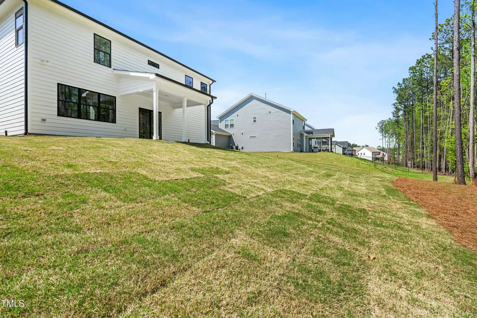 A large, gently sloping backyard with fresh sod behind a modern white house; other similar houses and tall pine trees are visible along the edge on a clear, sunny day.