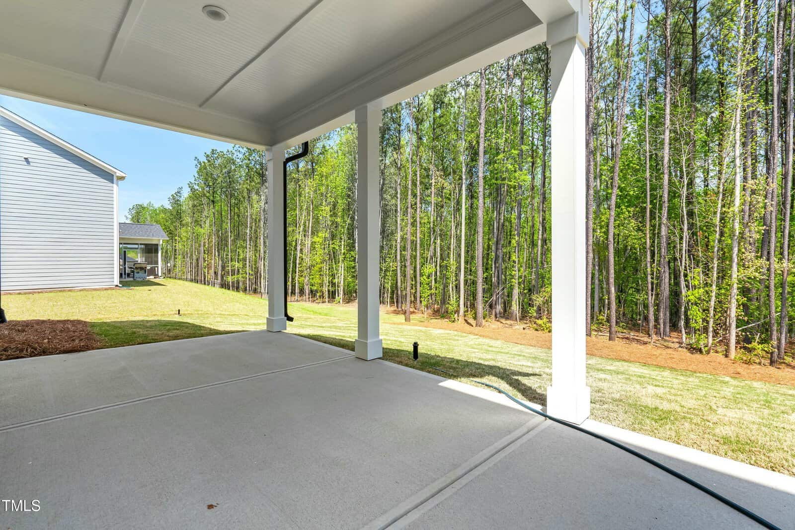 Covered patio with white columns and a concrete floor, overlooking a grassy yard bordered by tall, dense trees under a clear blue sky. A neighboring house is partially visible to the left.