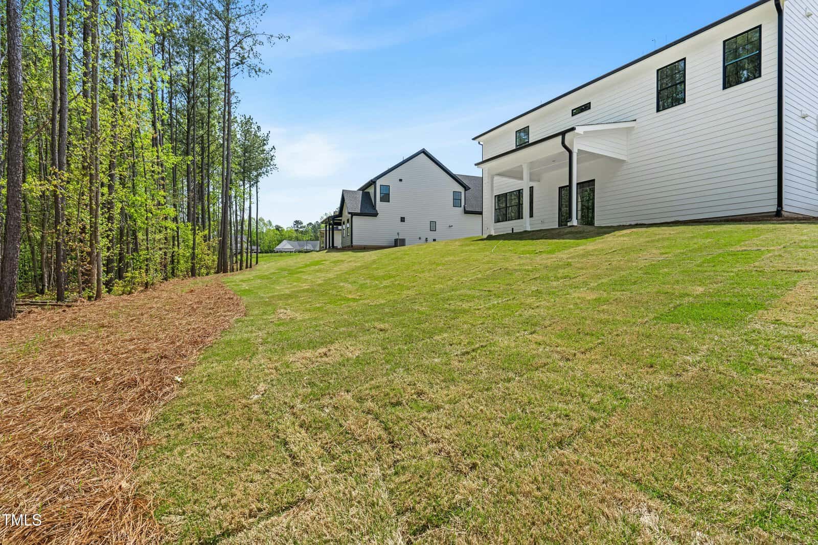 A grassy backyard slopes gently downward beside a white house with large windows and a covered patio; tall trees border the left side under a clear blue sky.