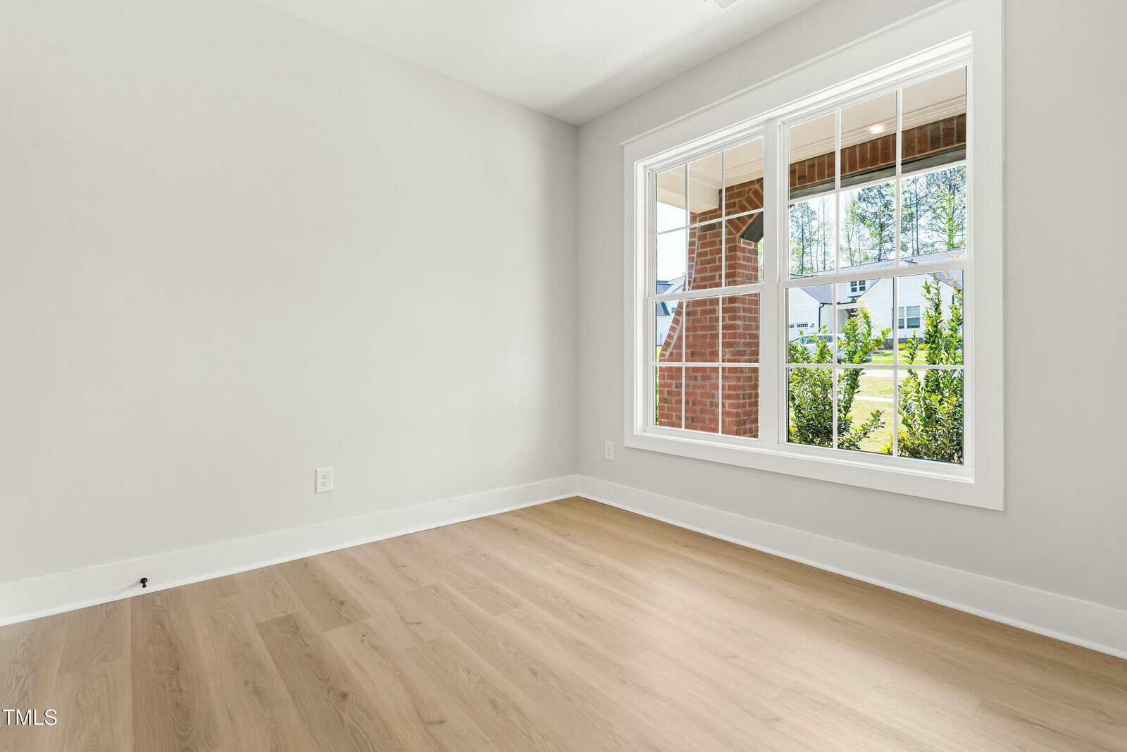 Bright, empty room with light wood floors, pale gray walls, and a large window overlooking a porch and green yard. The room is unfurnished and filled with natural light.