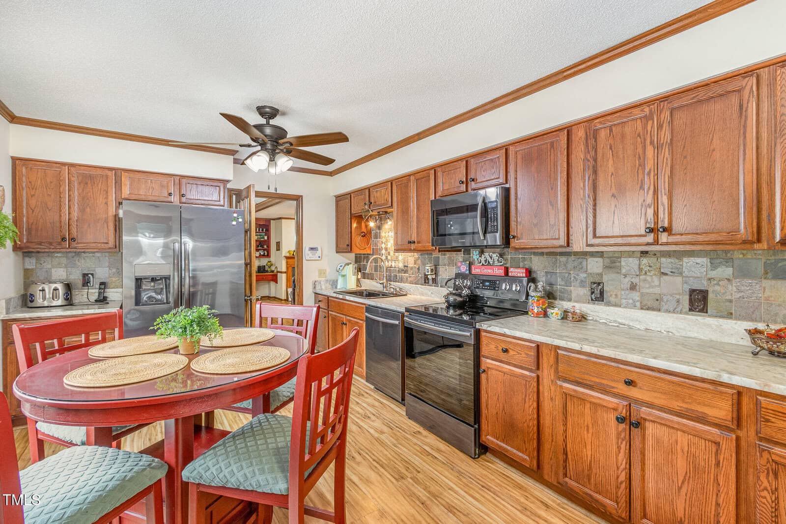 A kitchen with wood cabinets and a stone-tile backsplash. It features stainless steel appliances, including a double-door refrigerator and a black dishwasher. A round wooden table with chairs is set nearby, topped with place mats and a potted plant.