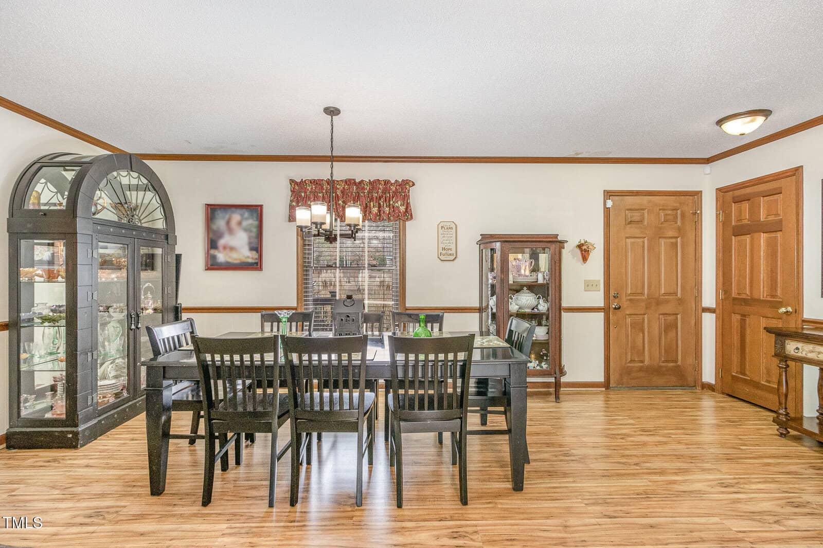 A dining room with a wooden table and eight matching chairs. On the left, a large cabinet with glass doors displays various items. A smaller cabinet is to the right. A window with a curtain sits in the middle. The floor is light wood.