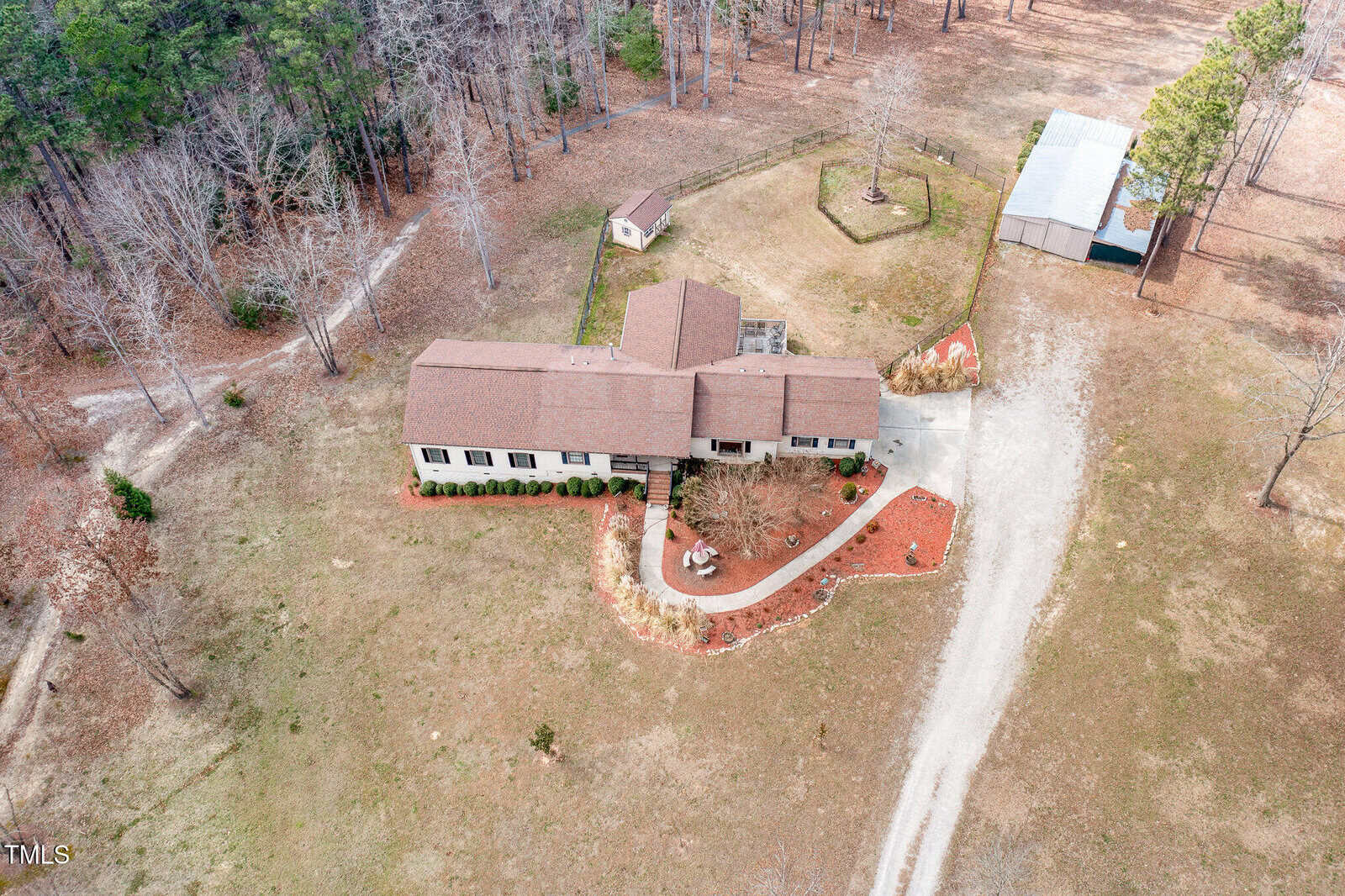 Aerial view of a suburban property with a single-story house, brown roof, and landscaped garden. A curved driveway leads to the entrance. There's a small outbuilding and a large rectangular shed on the right, surrounded by wooded areas.
