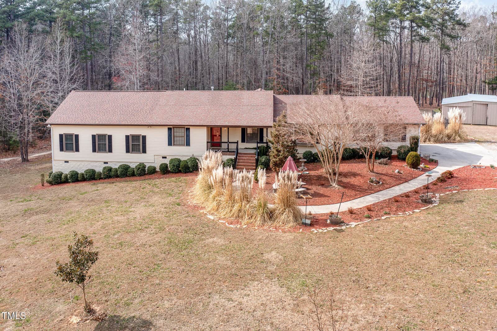 A single-story house at 89 Mount Pleasant Lane has a beige exterior and brown roof, nestled in a yard with minimal grass. The property features a paved walkway, small shrubs, and mulch landscaping, surrounded by tall trees stripped of leaves.