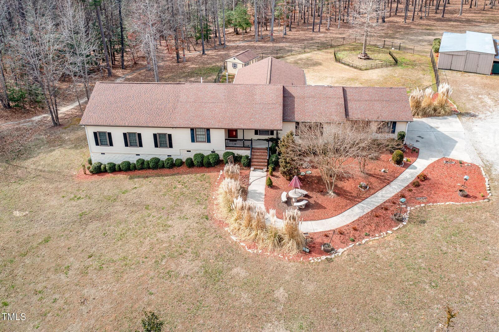 Aerial view of a single-story house with a brown roof, surrounded by a landscaped garden with red mulch, shrubs, and a paved walkway. The yard is expansive with dry grass, and a small structure is visible in the backyard near the trees.