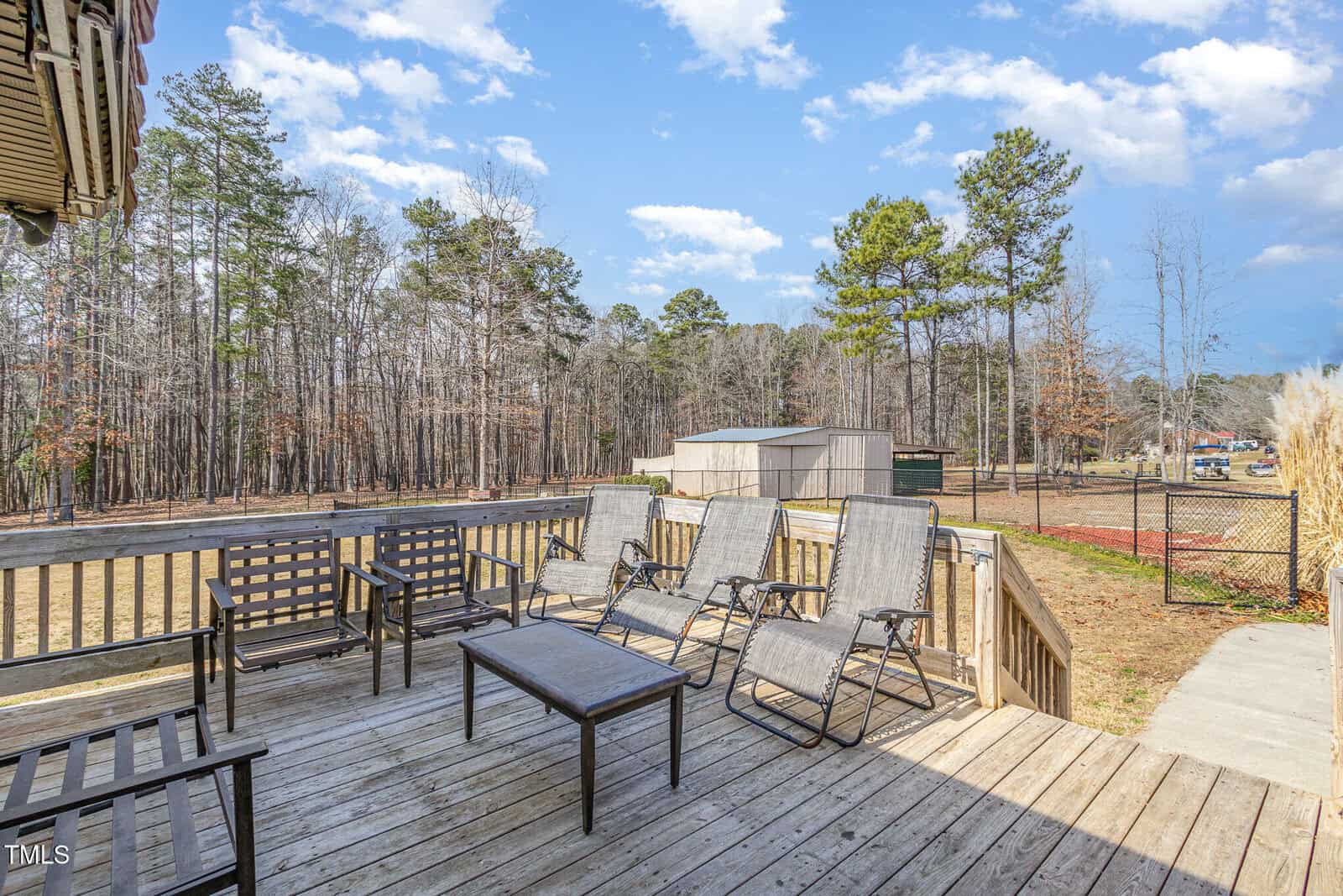 Wooden deck with six reclined lounge chairs, two black benches, and a central table. The deck overlooks a grassy yard with surrounding trees under a partly cloudy sky. In the background, there are storage sheds and a fenced area.