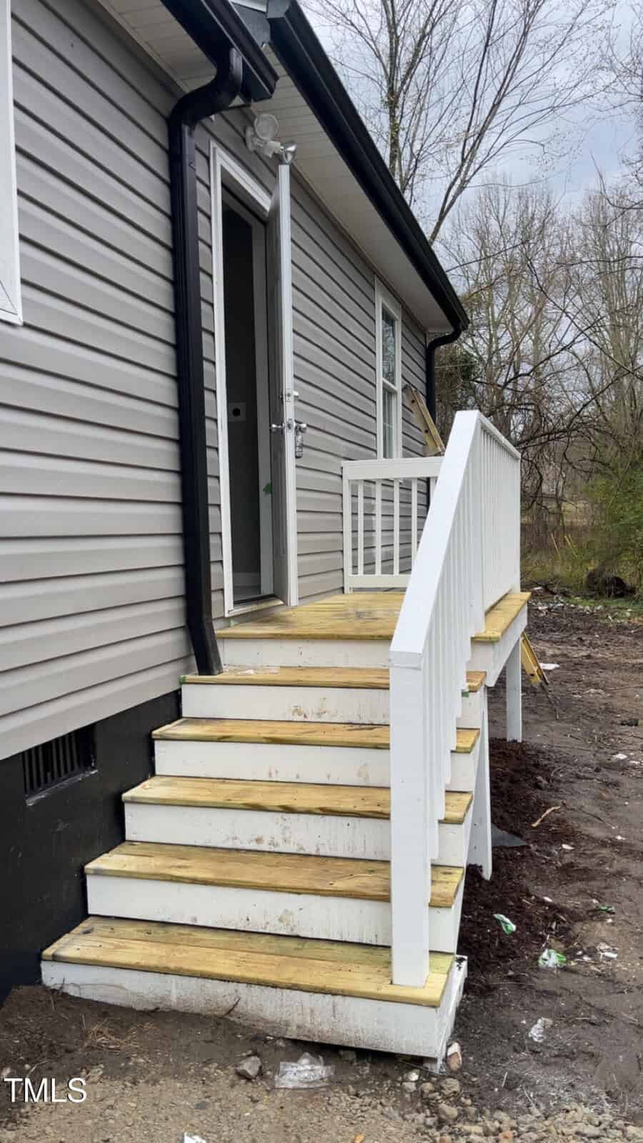 A small wooden staircase with white railings leads to the side entrance of a gray building. The door is open, revealing a glimpse of the interior. The surrounding area has bare ground and scattered debris, with trees in the background.