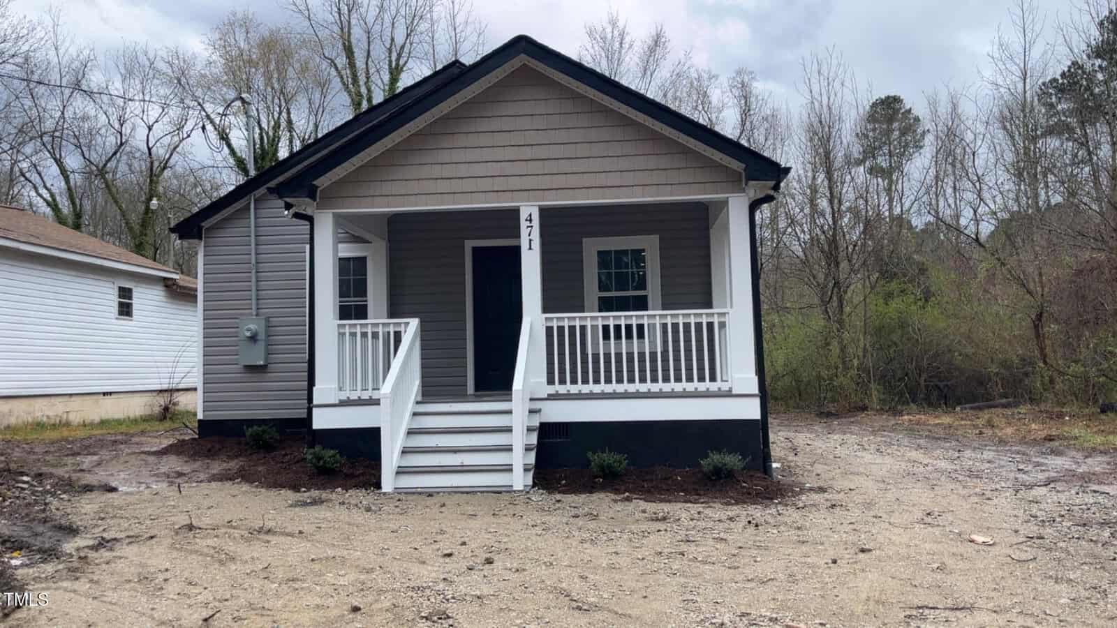 A small, single-story house with gray siding and a porch featuring white railing stands quietly at 471 Merriman Street. The yard is bare dirt with a few plants by the foundation. In the background, trees reach for cloudy skies in charming Henderson.