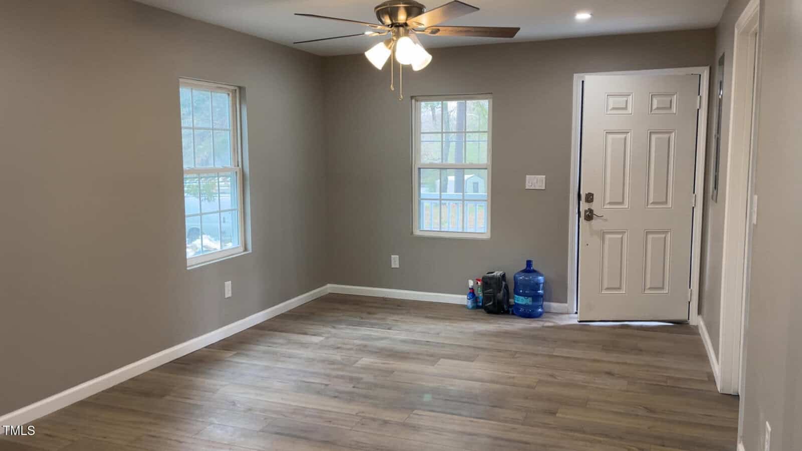 Empty room with light gray walls and wooden flooring. Features a ceiling fan with lights, two windows, and a closed white door. A filled blue water jug and some bags are placed near the door next to an electrical outlet.