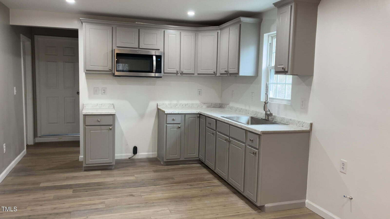 A modern kitchen with light gray cabinets, a marble countertop, and a stainless steel sink. There is a built-in microwave above the counter. The floor is wood, and there is a window above the sink letting in natural light.