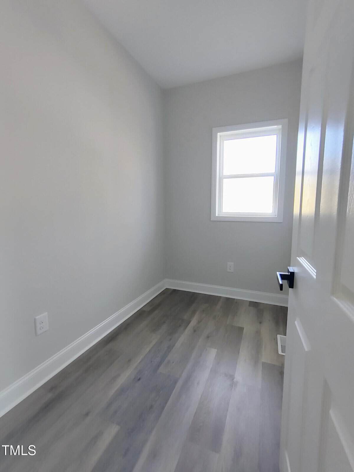 Small empty room with light gray walls, a wood-grain laminate floor, and a white trim window. The room is seen from an open white door with a black handle. No furniture is present.