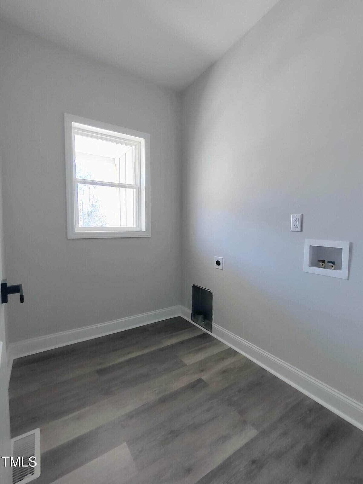 Small, empty room with light gray walls and vinyl plank flooring. A window on the left wall lets in natural light. There's a plumbing and electrical setup for a washer and dryer on the right wall.
