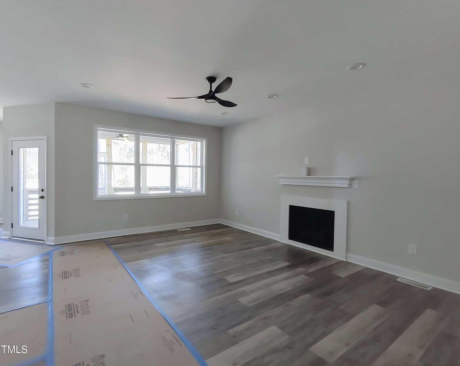 A vacant living room with light gray walls and wood laminate flooring. It features a black ceiling fan, a white fireplace with a mantel, and a large window. The floor is partially covered with protective paper near the entrance.