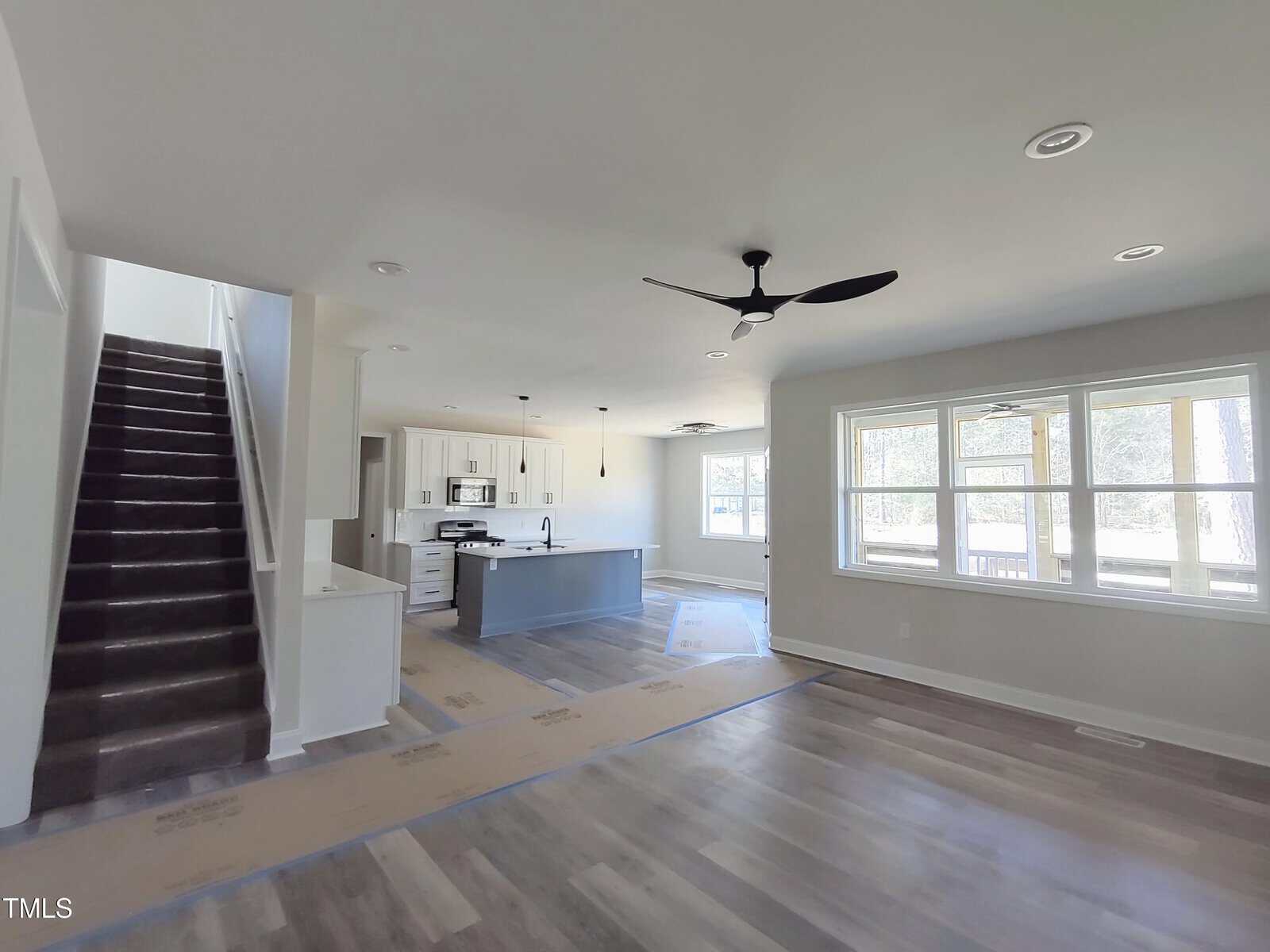 A modern, unfurnished open-plan living space featuring a kitchen with an island, stainless steel appliances, and pendant lights. The room has wooden floors, large windows, a ceiling fan, and a view of a staircase leading to the upper floor.