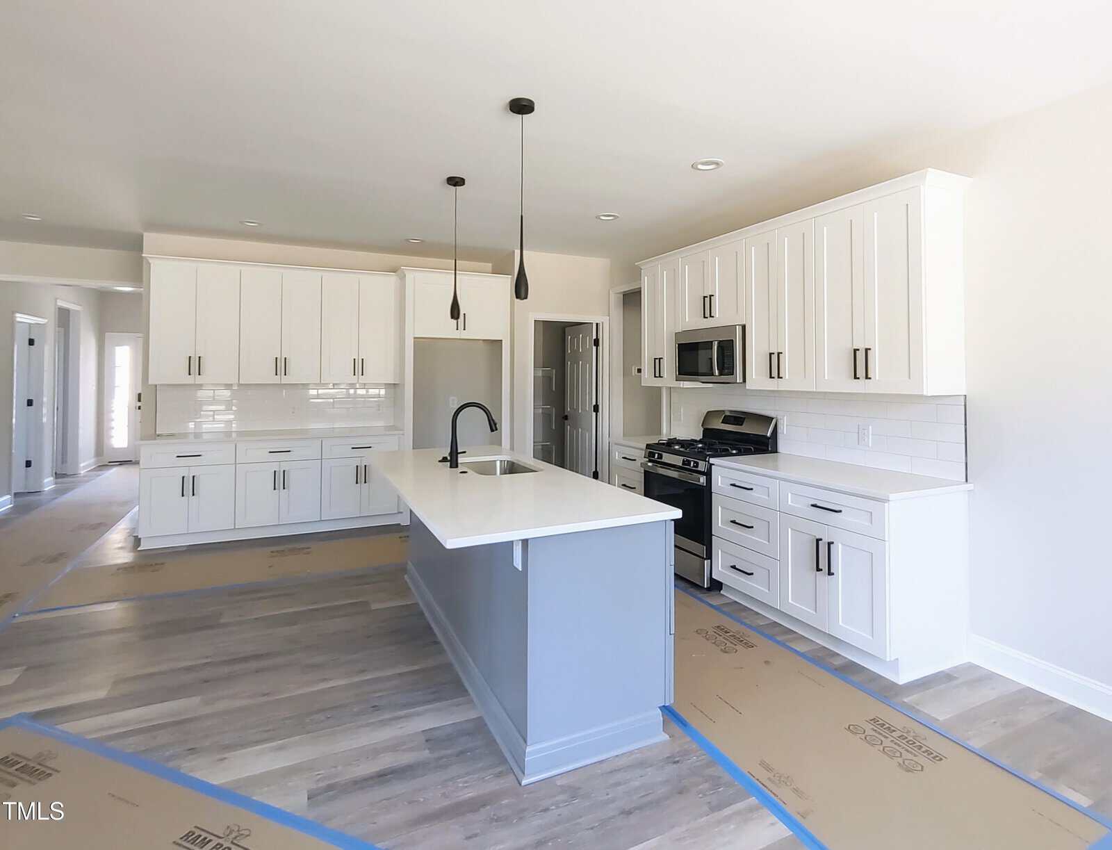 A modern kitchen with white cabinetry, a central island, and stainless steel appliances. The floor is light wood, and the countertops are white. Three black pendant lights hang above the island, and cardboard is laid out on the floors.