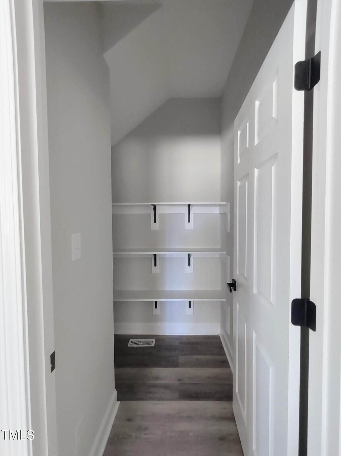 A small, empty walk-in closet with white walls and ceiling, featuring wooden flooring. Three white shelves with dark brackets are mounted on the back wall. The closet door is open, revealing a white frame and black hinges.