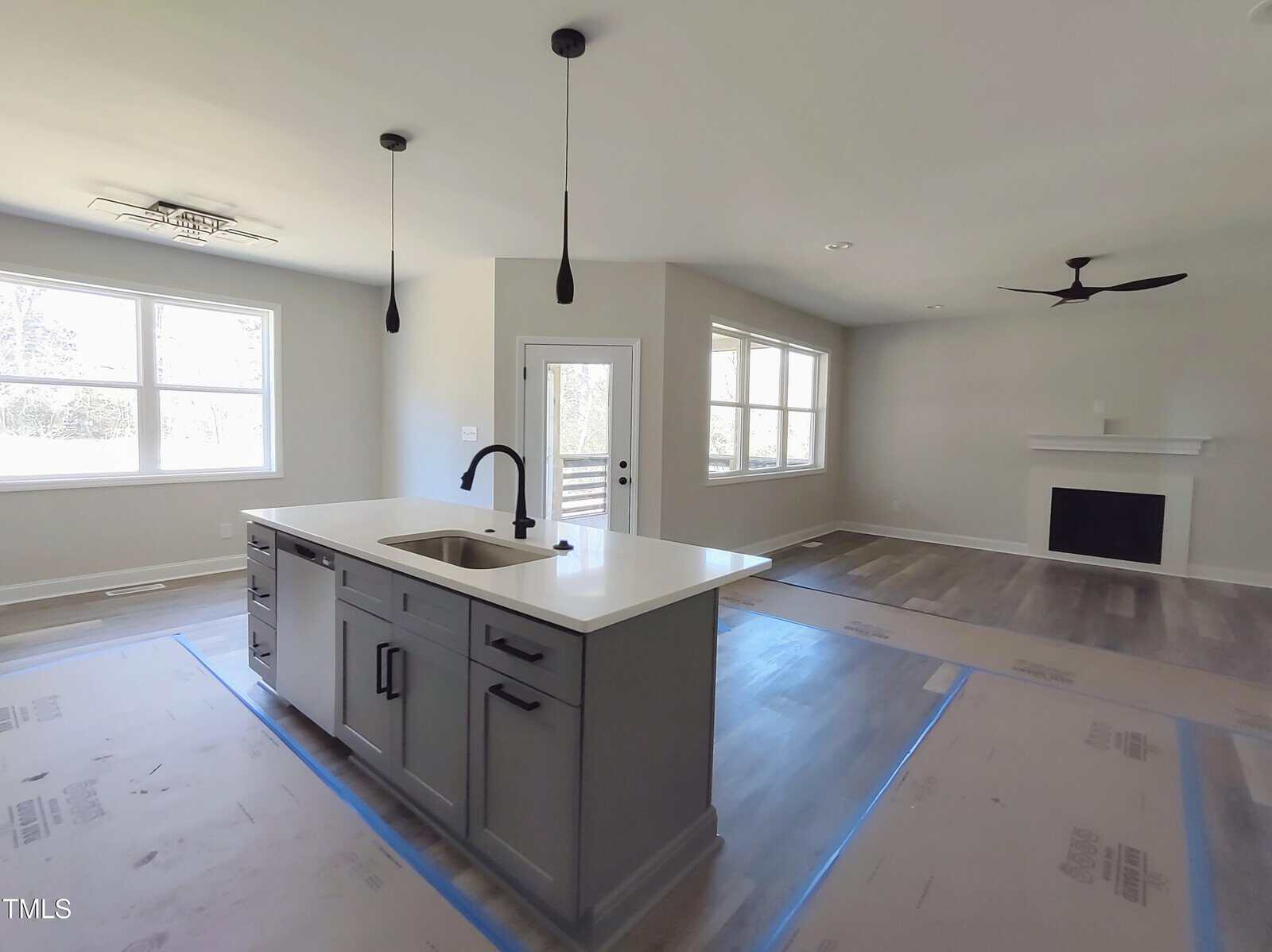 Modern open-concept kitchen and living room. The kitchen features a large island with a sink and pendant lights. The living area has large windows and a fireplace. Floors are covered for protection. Walls are painted light gray.