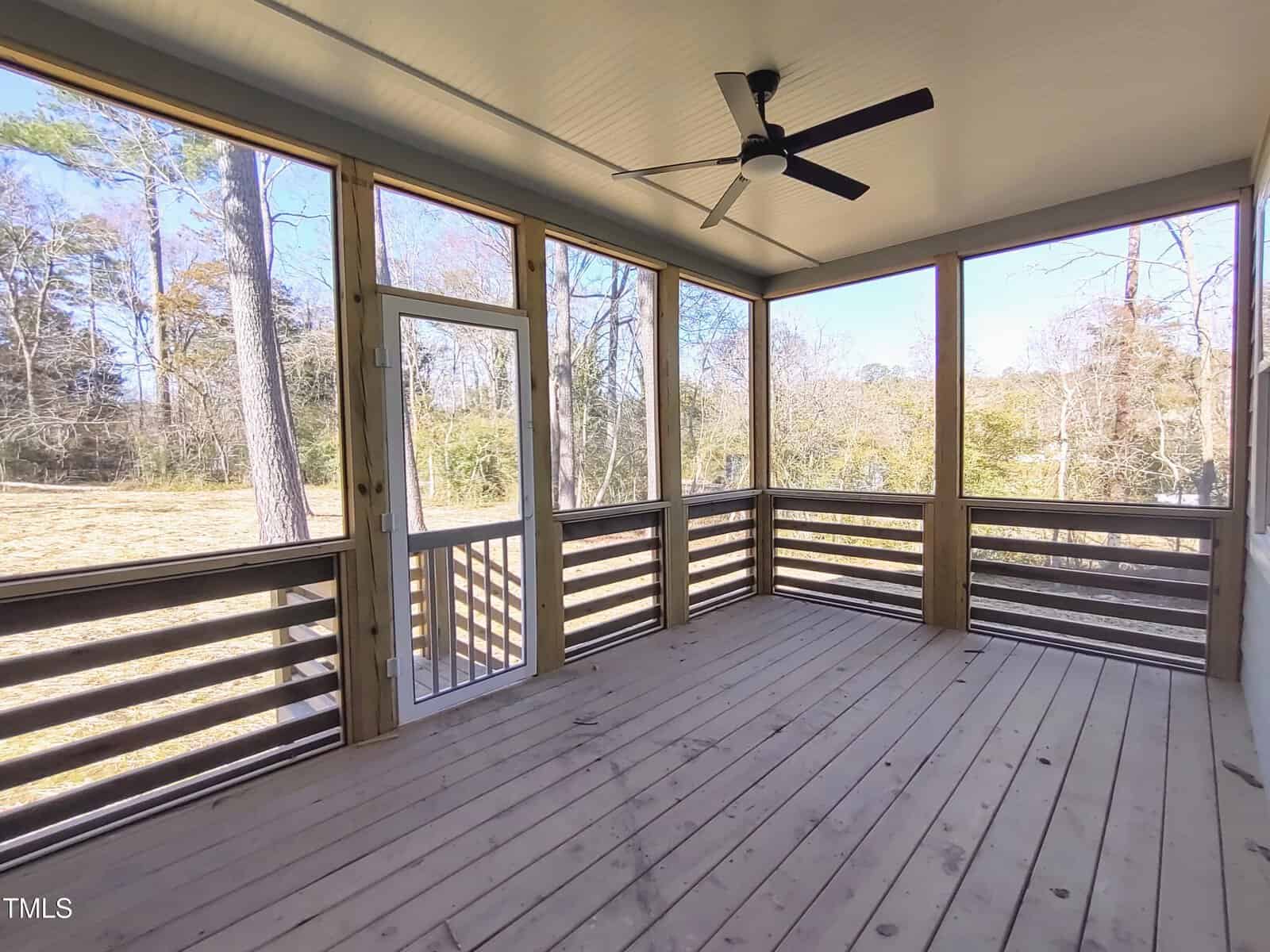 A screened porch with wooden flooring and railing, featuring a white ceiling with a black ceiling fan. The porch has large screened windows and a screen door, offering views of a grassy yard and trees.