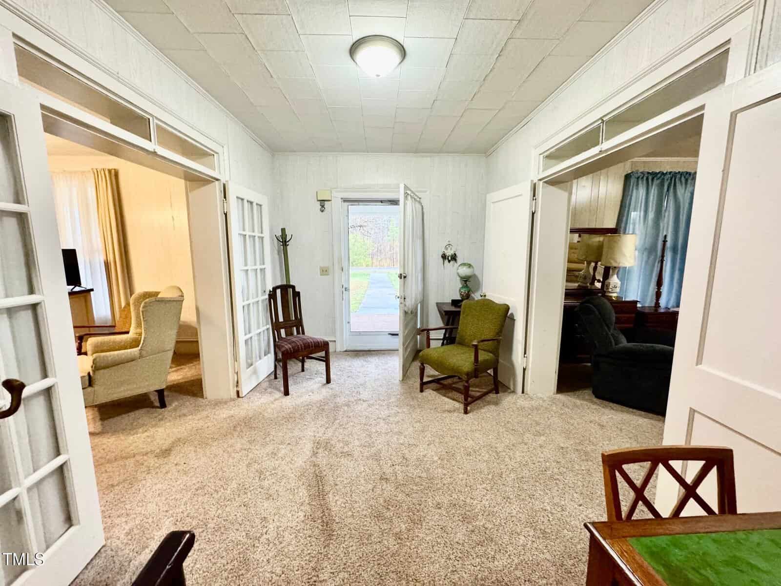 A living room with light-colored carpet and white walls features an armchair and wooden chairs. French doors lead to a hallway, with a view of a glass door and outdoor scene. A table with a lamp is visible in the background.