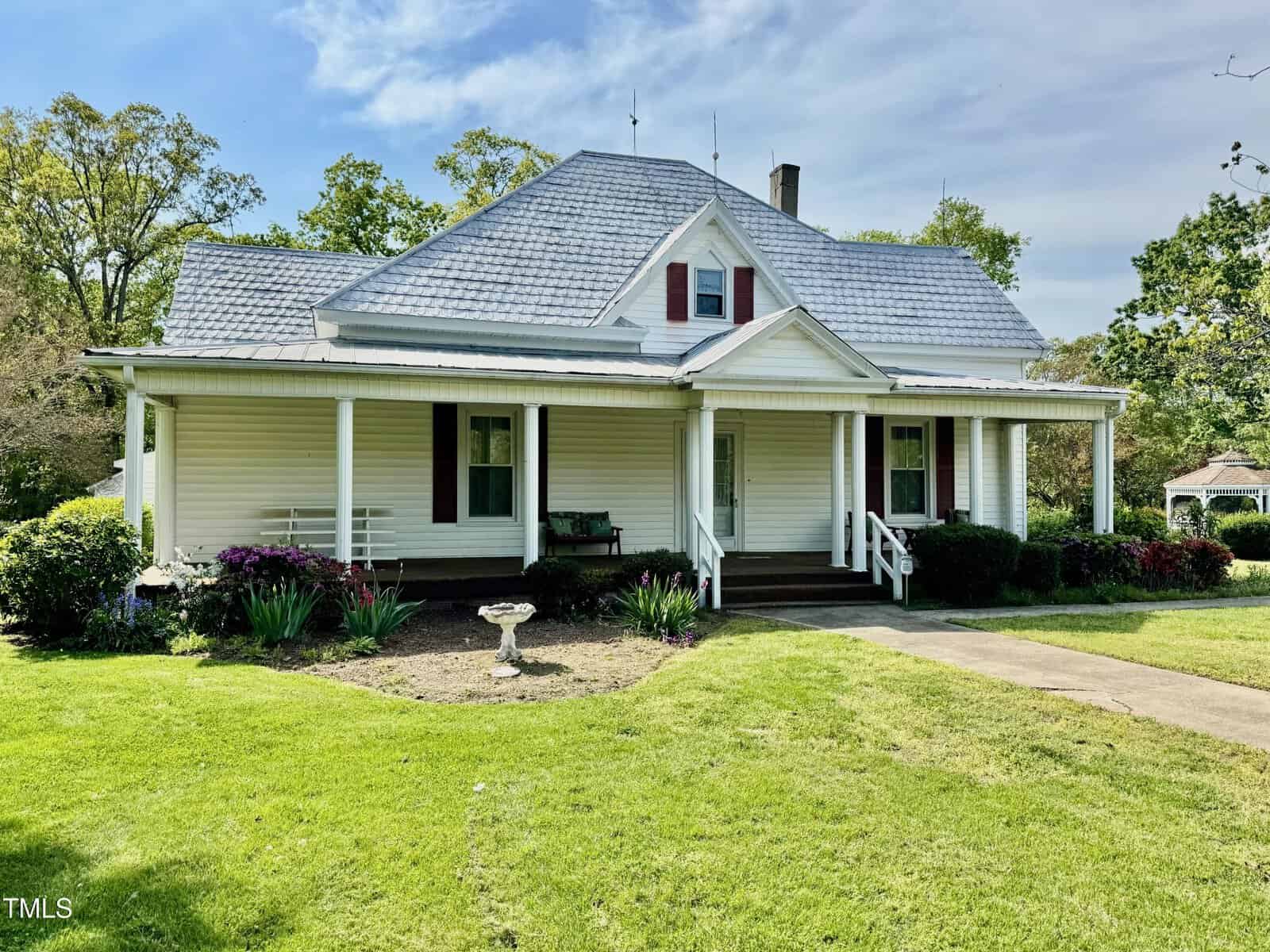 Nestled at 4715 Jacksontown Road in Manson, this white house features a gray roof and a charming covered front porch. The well-maintained yard boasts a lush lawn, a small garden, and a birdbath near the walkway, with trees and shrubs surrounding the property and a gazebo gracing the background.
