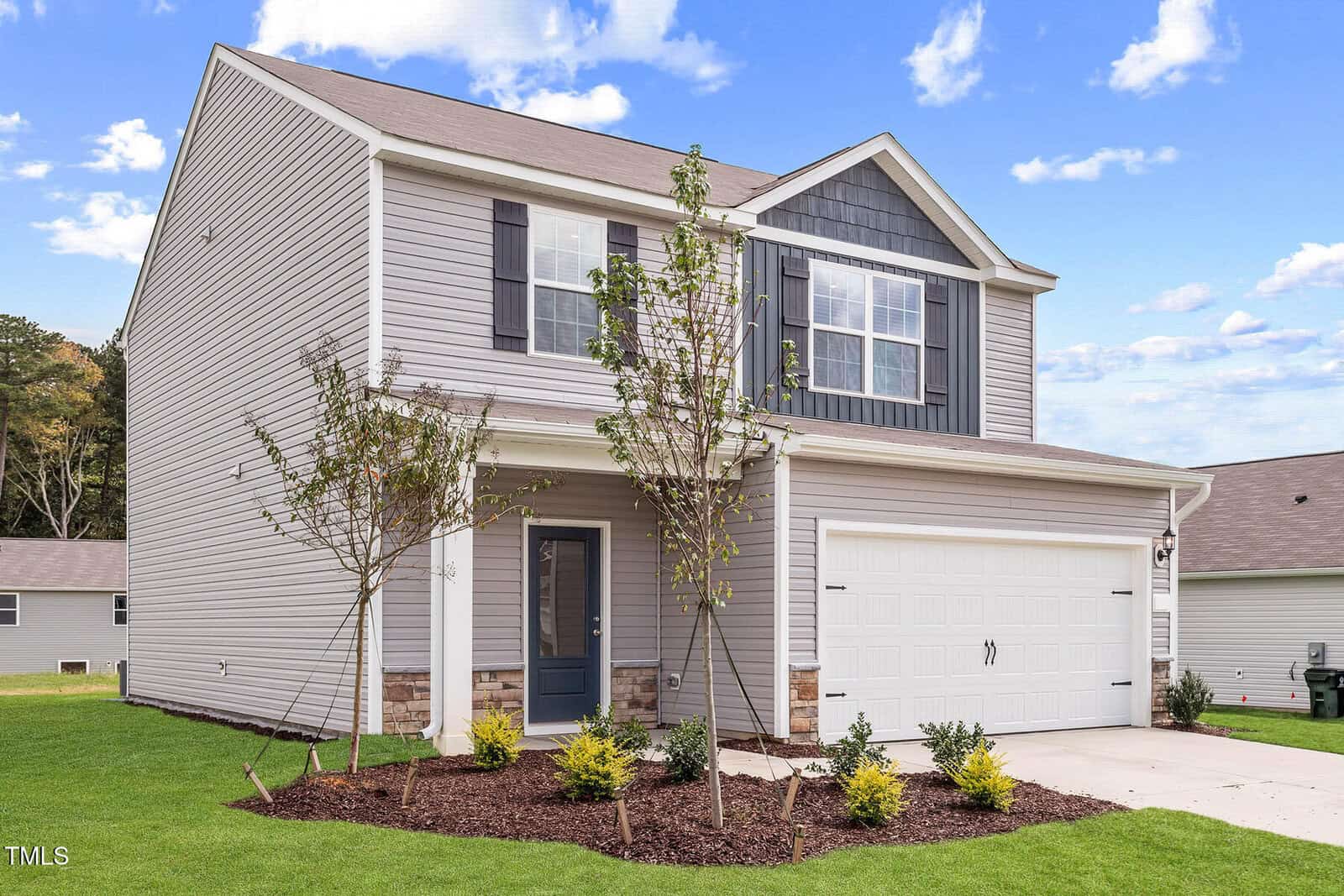 A charming two-story house on 110 Rhino Bend features gray siding and a dark blue front door. With a two-car garage and light brick accents, it exudes elegance. Nestled in Oxford, the small front yard boasts young trees and shrubs under a clear sky dotted with a few clouds.