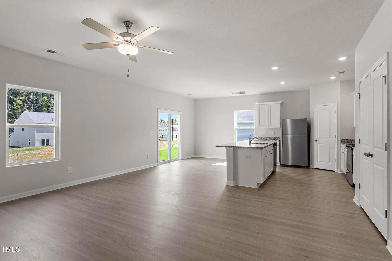 Open concept kitchen and living area with light wood flooring. The kitchen features white cabinets, a silver refrigerator, and an island. A ceiling fan is in the living area. Large windows and a sliding door lead to a grassy backyard.