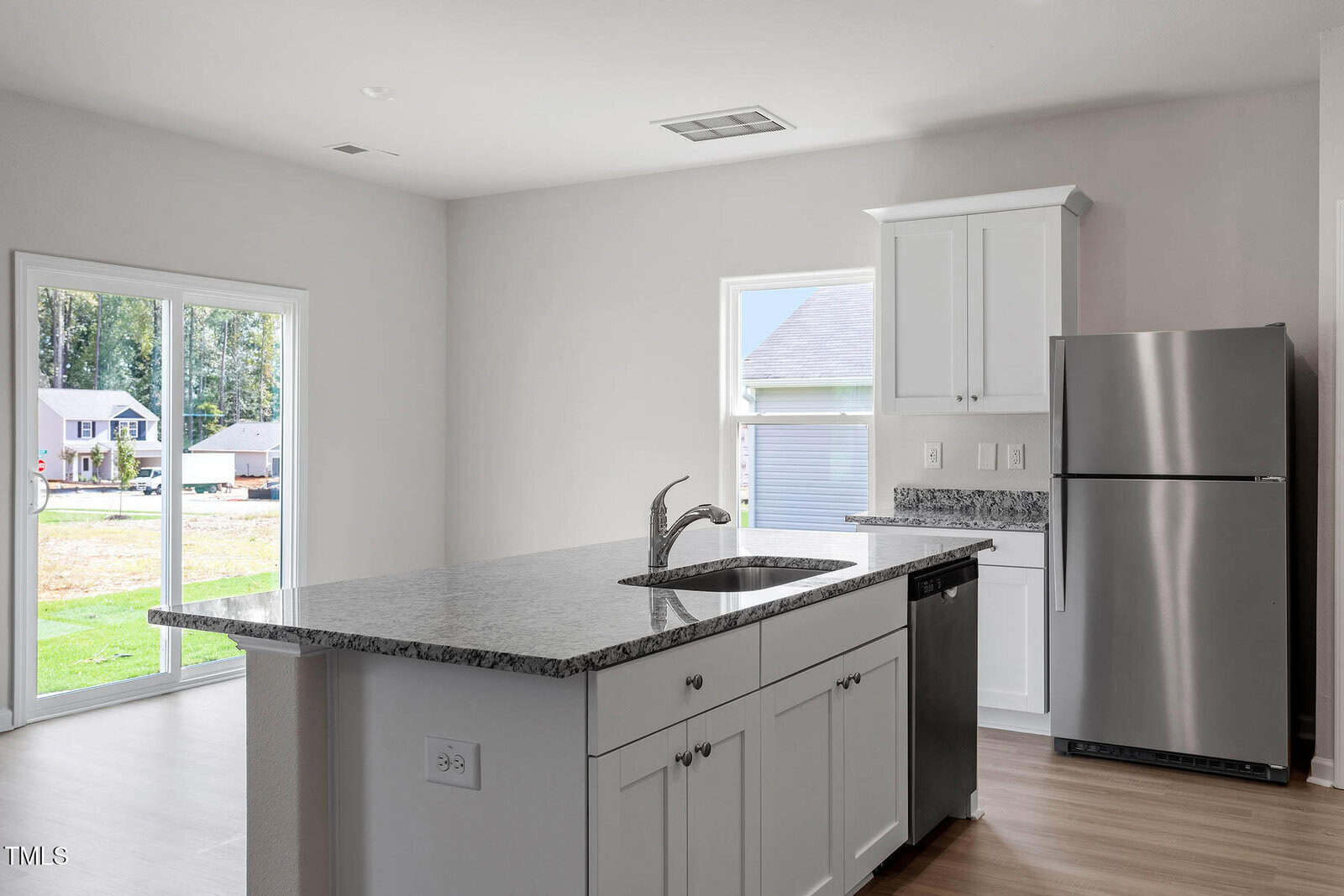 Modern kitchen with a large island featuring a granite countertop and a built-in sink. White cabinets line the walls, and stainless steel appliances, including a refrigerator and dishwasher, are visible. Sliding glass doors lead to the outside.