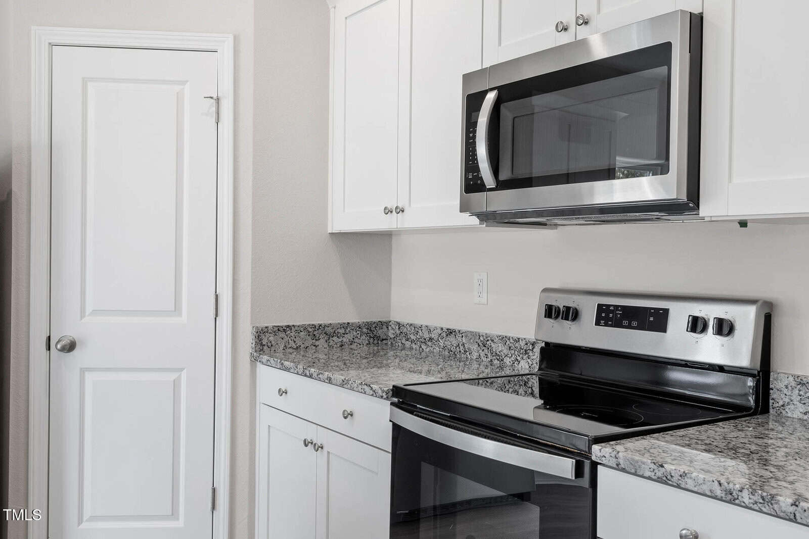 Modern kitchen with white cabinets, gray granite countertops, and a stainless steel oven with a matching microwave above. A white door is to the left. The walls are light beige.
