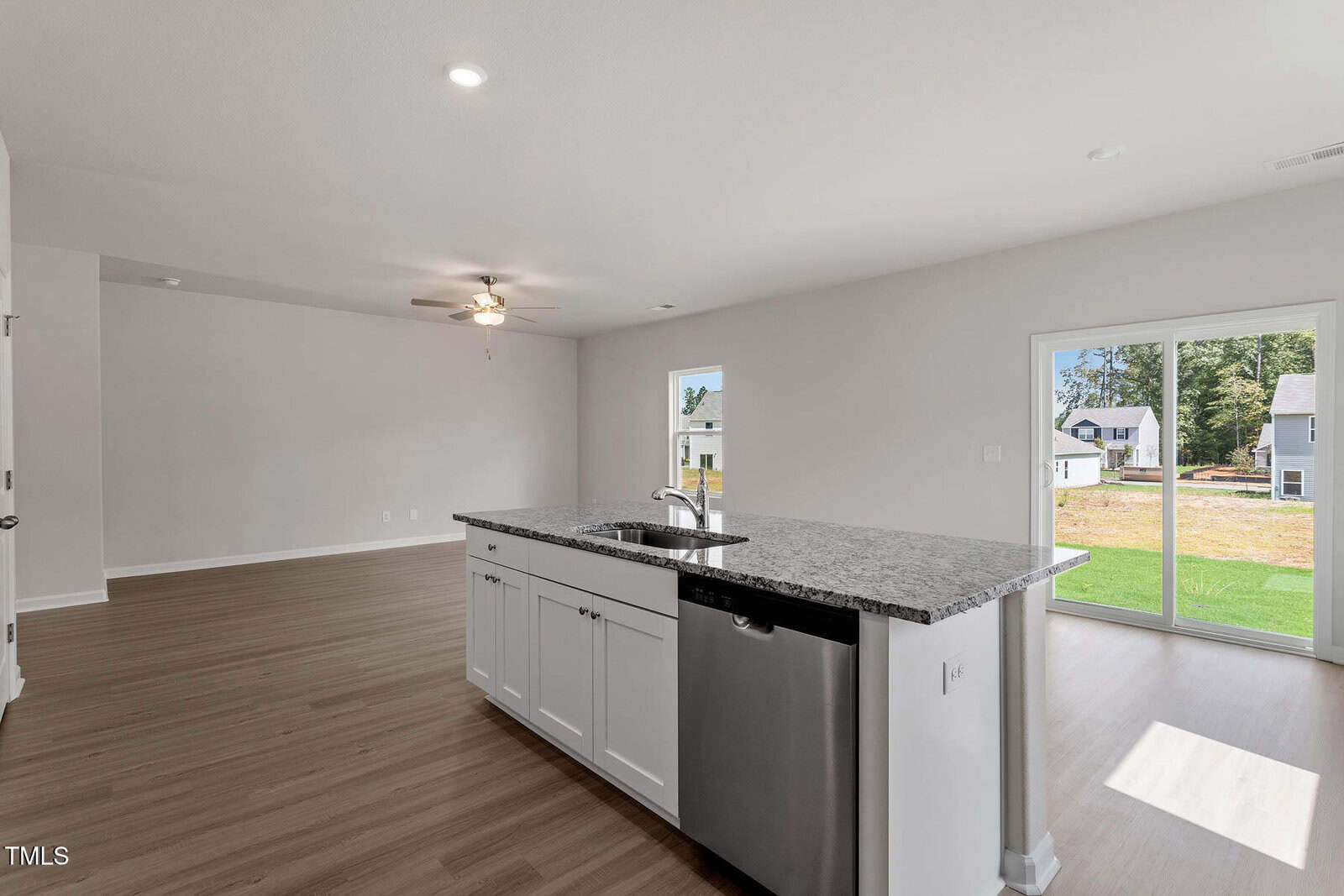 A modern kitchen and living area with light wood flooring. The kitchen features a center island with a granite countertop and a built-in stainless steel dishwasher. A ceiling fan hangs in the living space. Large windows offer a view of a grassy backyard.