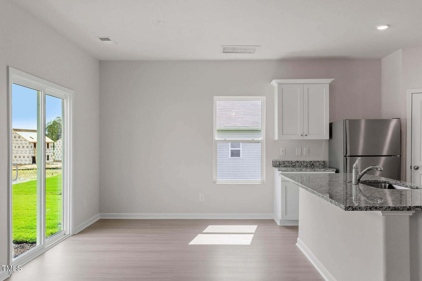A bright kitchen and dining area with light wood flooring. Features include a granite countertop, stainless steel refrigerator, white cabinets, and a double sink. Sliding glass doors lead to a grassy outdoor area. Natural light streams through a window.