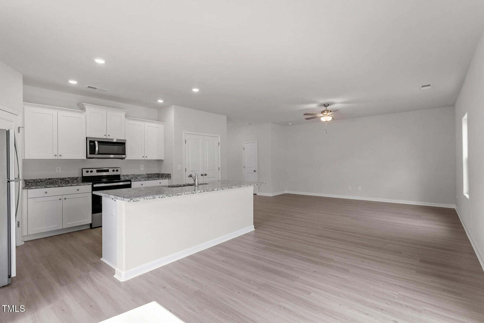A spacious, unfurnished kitchen and living area with light wood flooring. The kitchen features white cabinets, a granite countertop island, and stainless steel appliances. A ceiling fan is in the living space, and natural light comes through the window.