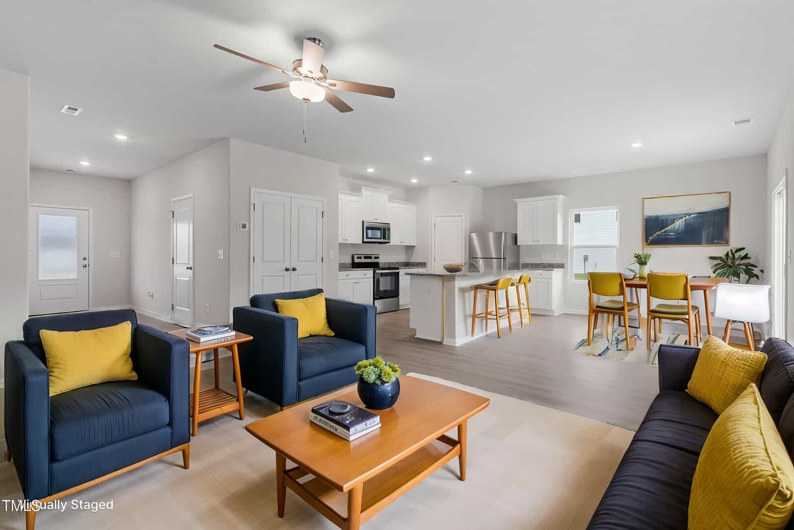 A modern, open-concept living space featuring a kitchen, dining area, and sitting area. The kitchen has white cabinets and stainless steel appliances. The sitting area includes dark chairs, a yellow-accented sofa, and a wooden coffee table.