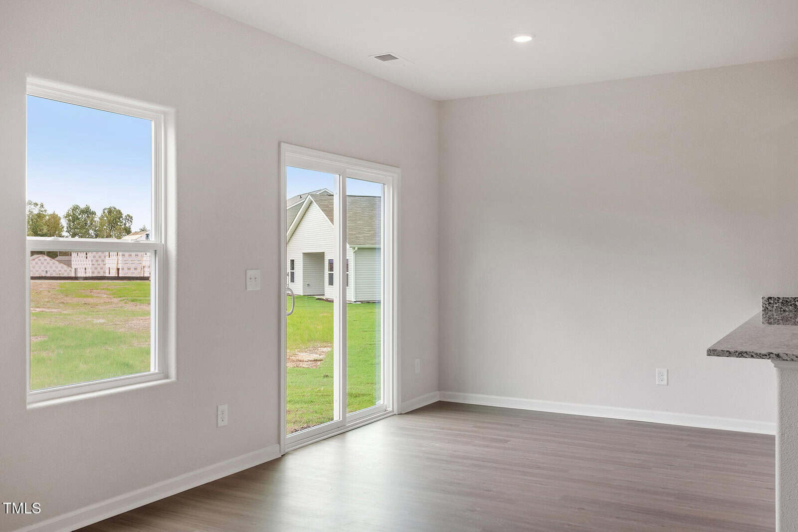 A bright, empty room with light-colored walls and wooden flooring. It has a large window and a glass sliding door leading outside. The room offers a view of a grassy area and a neighboring house in an underdeveloped area.
