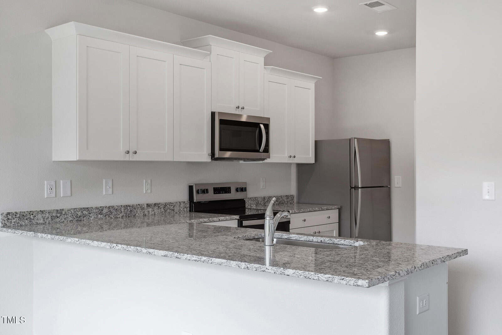 A modern kitchen with white cabinets, stainless steel appliances, and a granite countertop. There is a sink and a stove on the counter, a microwave above the stove, and a refrigerator in the background. The walls and ceiling are light-colored.