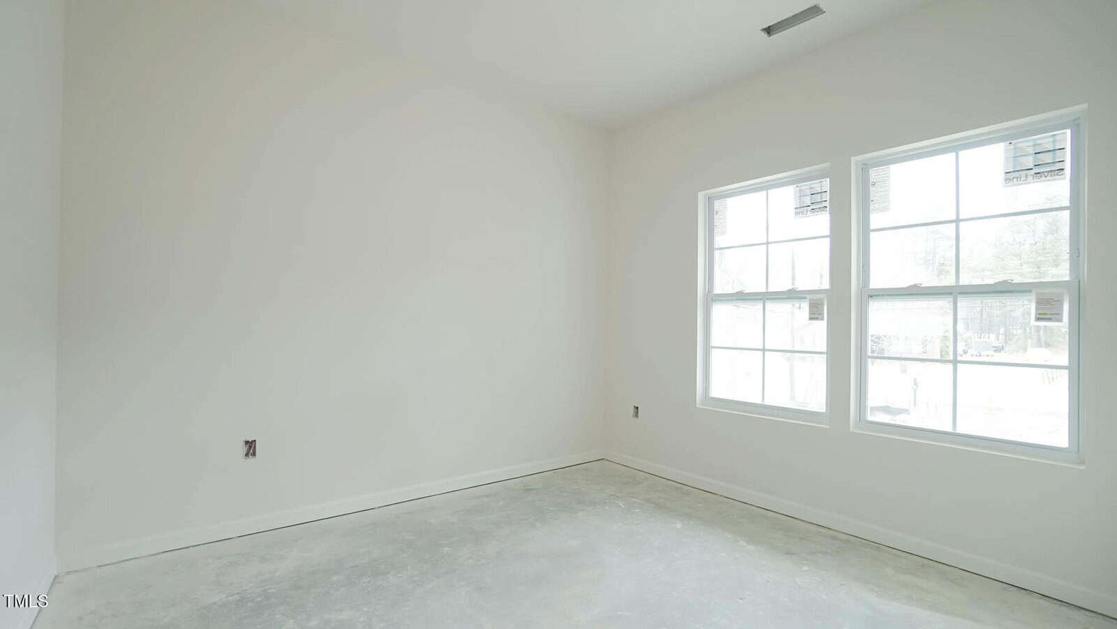 An empty, unfurnished room with unpainted walls and a concrete floor. Two large windows on the right side let in natural light. The space is under construction or renovation, with outlets and unfinished details visible.