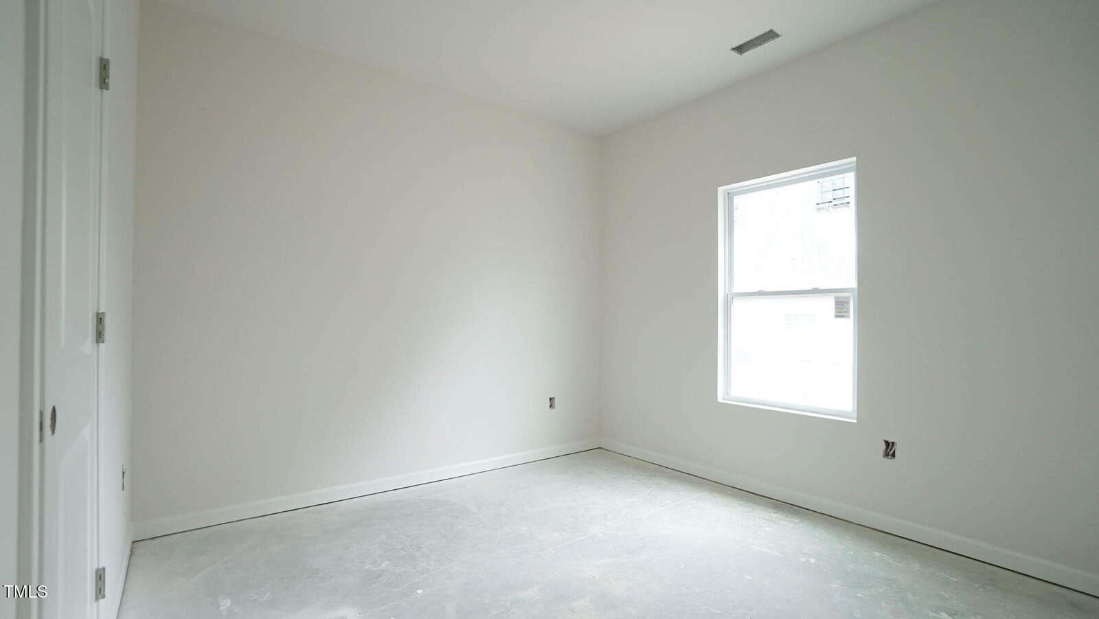 An empty room with white walls and a concrete floor. There is a single window on the right wall allowing natural light in. The room appears unfinished, with visible outlets and no furnishings.