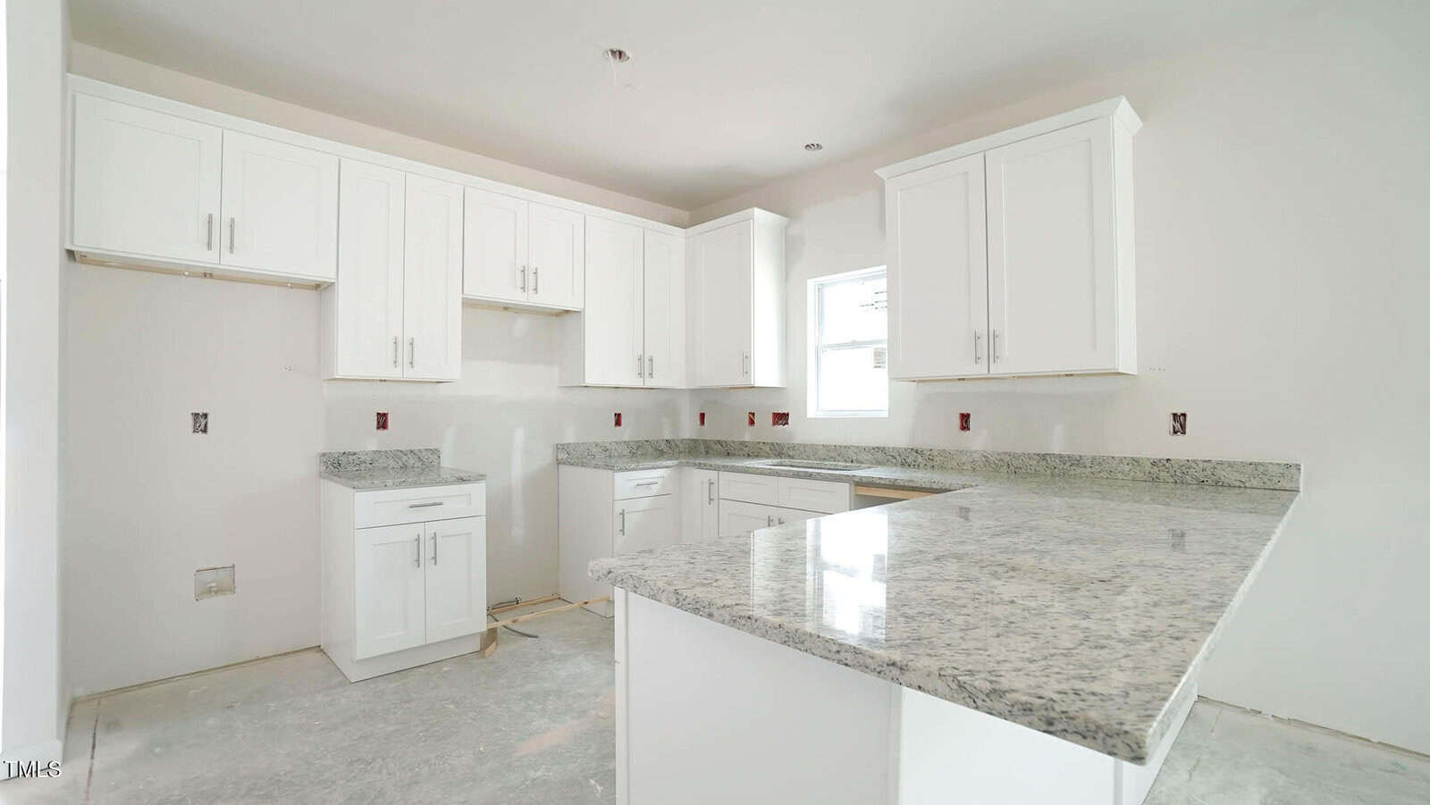 A kitchen under construction features white cabinets, gray granite countertops, and a light gray floor. The walls are painted white, and there is a window above the sink area. Electrical outlets are visible, but the appliances are not yet installed.