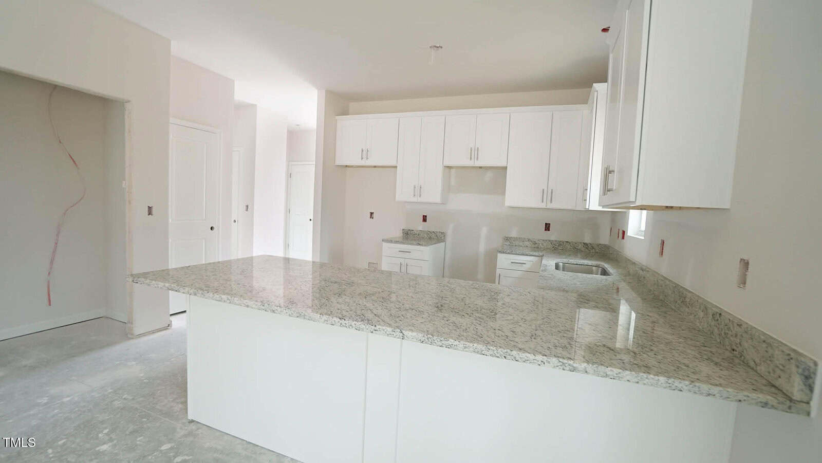 A modern kitchen with light-colored granite countertops, white cabinetry, and unfinished flooring. A window above the sink provides natural light. Walls are unpainted with visible outlets and light fixture placements.